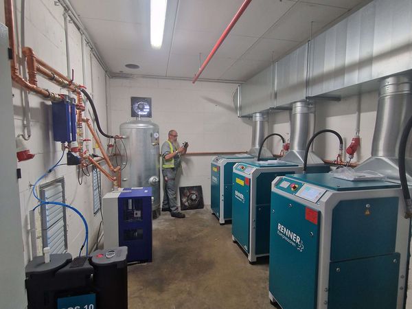 Technikraum mit Kompressoren und Leitungen – Mehrere Kompressoren mit komplexem Rohrsystem und einem Techniker bei der Wartung